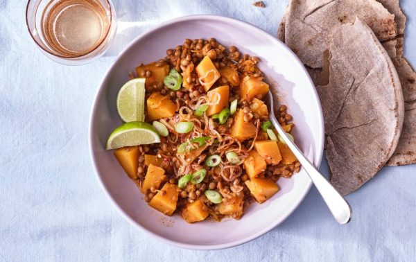 Een kom linzenstoofpot met stukjes gele pompoen, gegarneerd met gesneden groene uien en gebakken sjalotten, geserveerd met partjes limoen. Een glas lichtgekleurde drank en stukjes platbrood staan naast de kom op een lichtblauw tafelkleed.