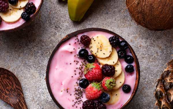 Naast bananen, een kokosnoot, een ananas en een houten lepel op een stenen oppervlak staat een smoothiebowl met daarop plakjes banaan, aardbeien, bramen, bosbessen en chiazaadjes.