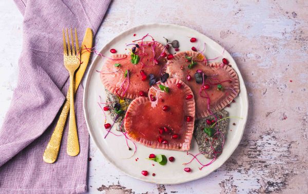 Op een wit bord ligt hartvormige roze ravioli, gegarneerd met microgroenten en granaatappelpitjes. Goud bestek rust op een lavendelkleurig servet ernaast. De achtergrond is een licht, gestructureerd oppervlak.