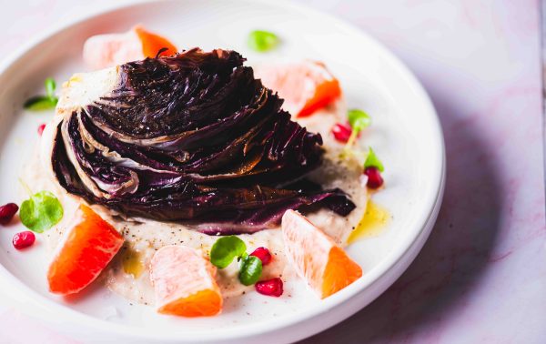 Op een bord ligt een partje gegrilde radicchio op een romige saus, gegarneerd met sinaasappelpartjes, granaatappelpitjes en kleine groene blaadjes. Het gerecht is kunstig gerangschikt op een wit bord.