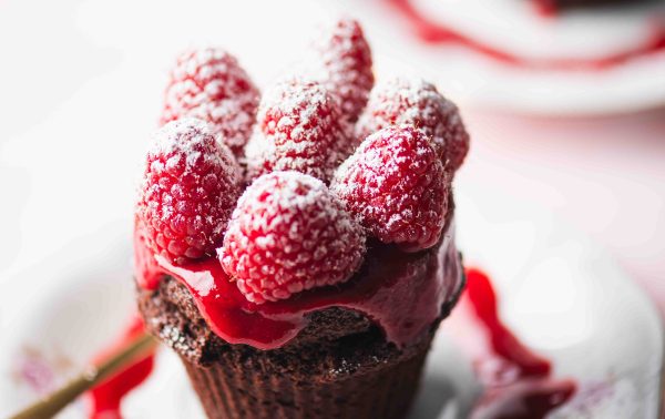 Een chocoladecupcake met daarop verse frambozen, frambozensaus en poedersuiker ligt op een wit bord, waar de saus kunstig omheen is gedruppeld.