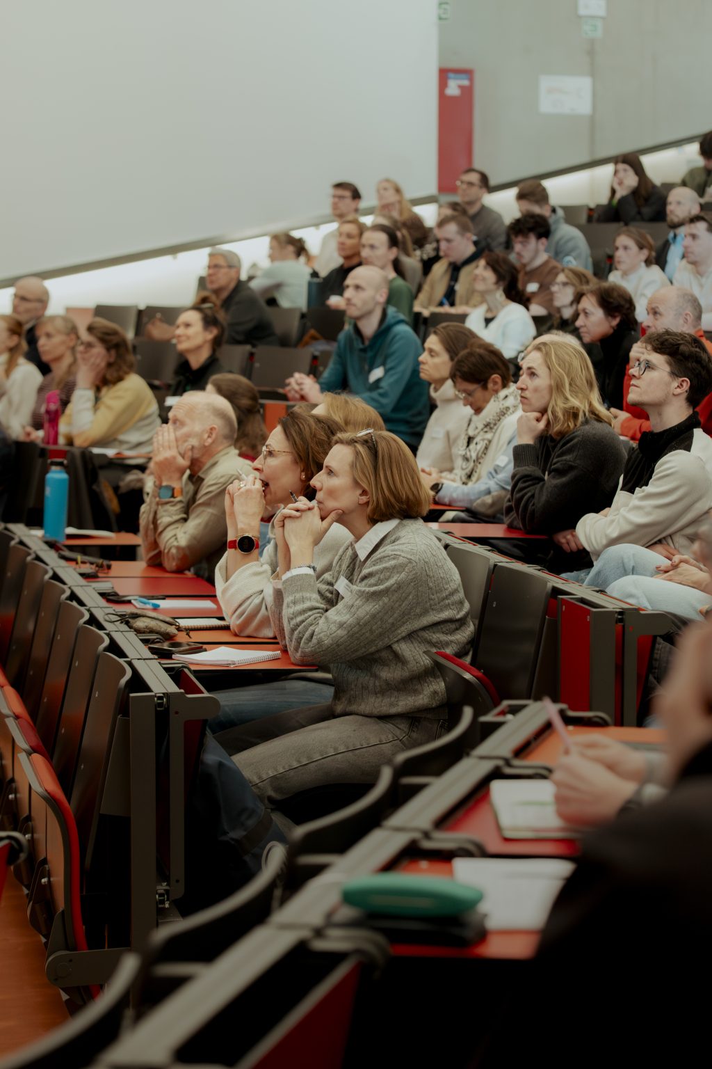Een groep mensen zit in rijen stoelen in een collegezaal, aandachtig te luisteren en aantekeningen te maken tijdens een presentatie of les.