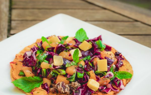 Een flatbread belegd met in blokjes gesneden groenten, groenten en verse munt wordt geserveerd op een vierkant wit bord op een houten tafel.