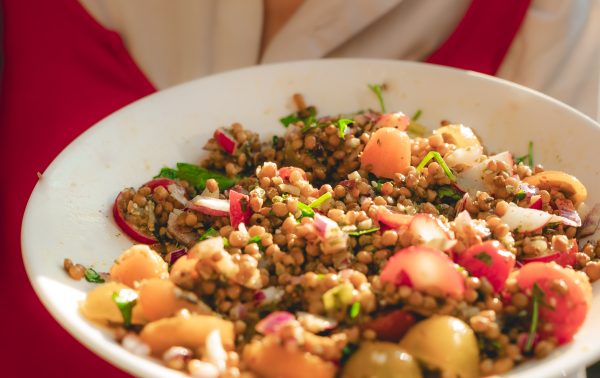 Persoon die een kom gemengde linzen en groentesalade vasthoudt en een lepel naar zijn mond tilt.