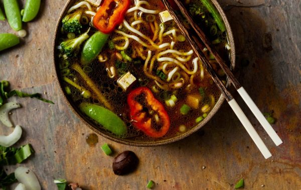 Kom noedelsoep met tofu, kikkererwten, rode paprika en sjalotten, met eetstokjes erop; verse groenten en kruiden verspreid op een rustiek houten oppervlak.