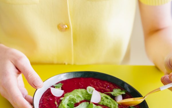 Een persoon in een geel shirt houdt een kom rode soep vast met daarop groene saus, plakjes radijs en koriander, met behulp van een gouden lepel op een gele tafel.