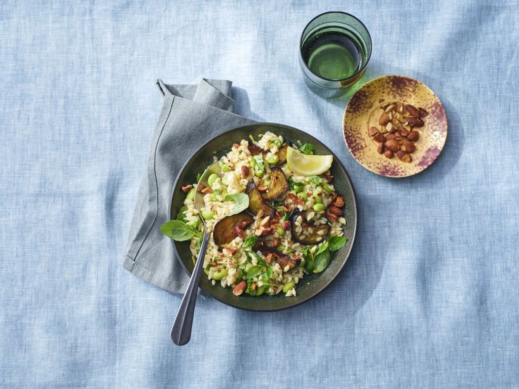 Een bord rijst met gegrilde aubergine, kruiden, partjes citroen en amandelen, vergezeld van een glas water en een schaaltje amandelen op een blauw tafelkleed.