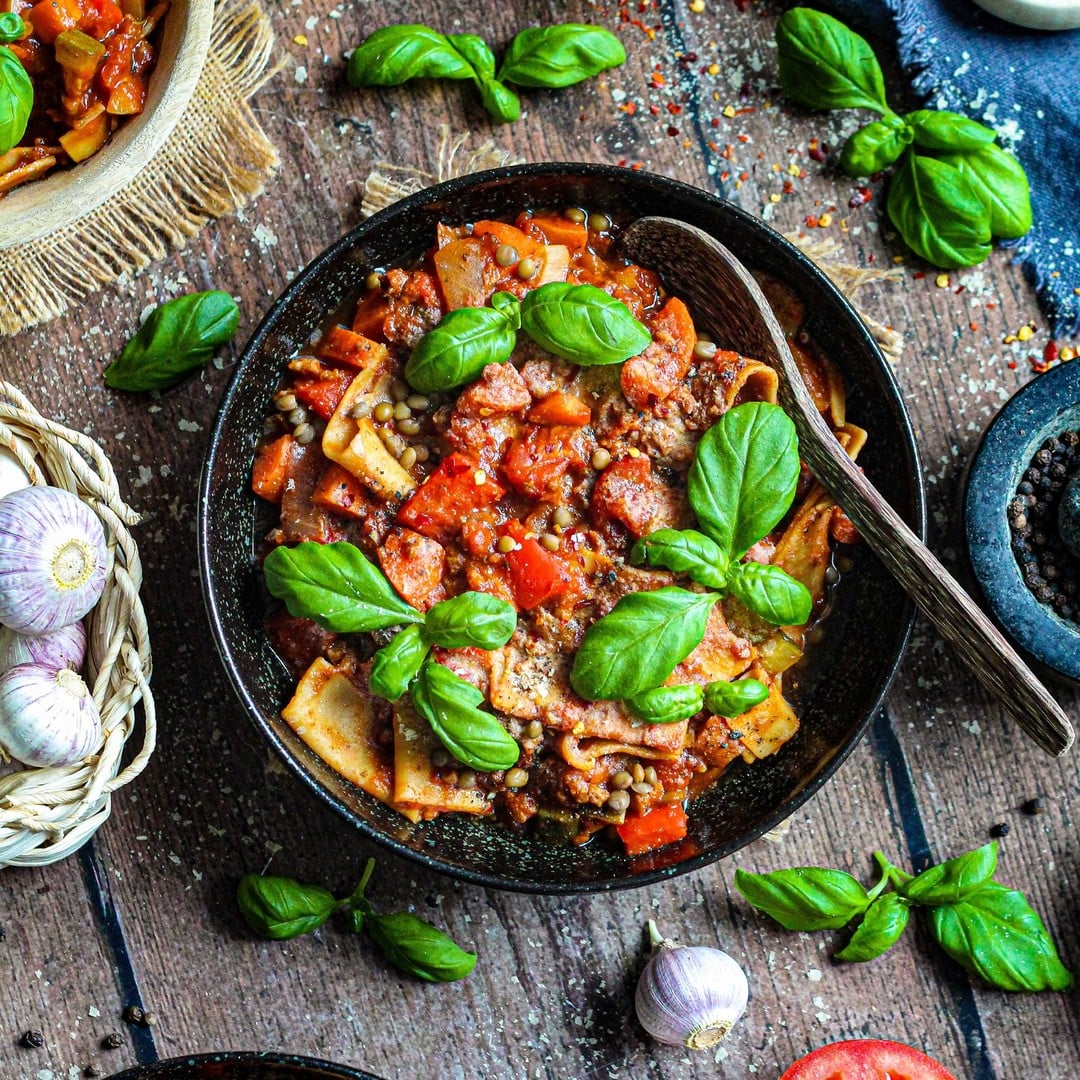 Een kom pasta met tomatensaus, gehakt en basilicumblaadjes, omringd door verse ingrediënten zoals knoflook, tomaten, peperkorrels en een selectie groenten uit de krabbelbak, geserveerd op een rustieke houten tafel.