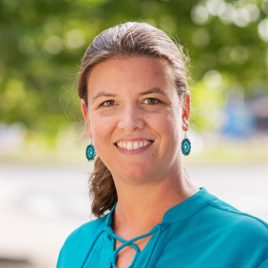 Vrouw met bruin haar in een paardenstaart, met turquoise oorbellen en een turquoise blouse, die buiten glimlacht tegen een wazige groene achtergrond.