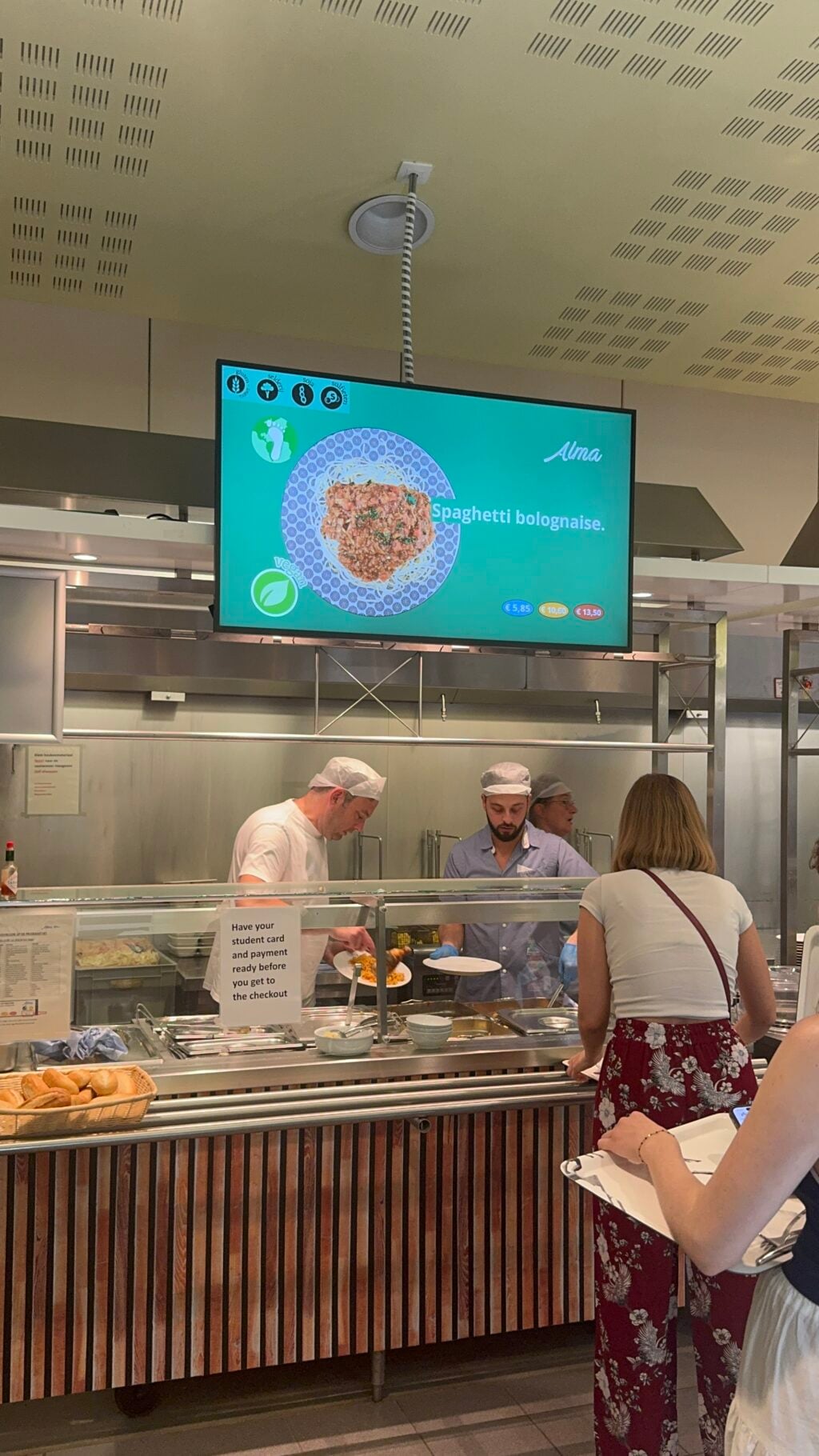 Twee cafetaria-medewerkers serveren eten aan mensen die in de rij staan, onder een digitaal menu waarop spaghetti bolognese staat vermeld, terwijl het campusaanbod van de KULeuven hongerige studenten blijft imponeren.