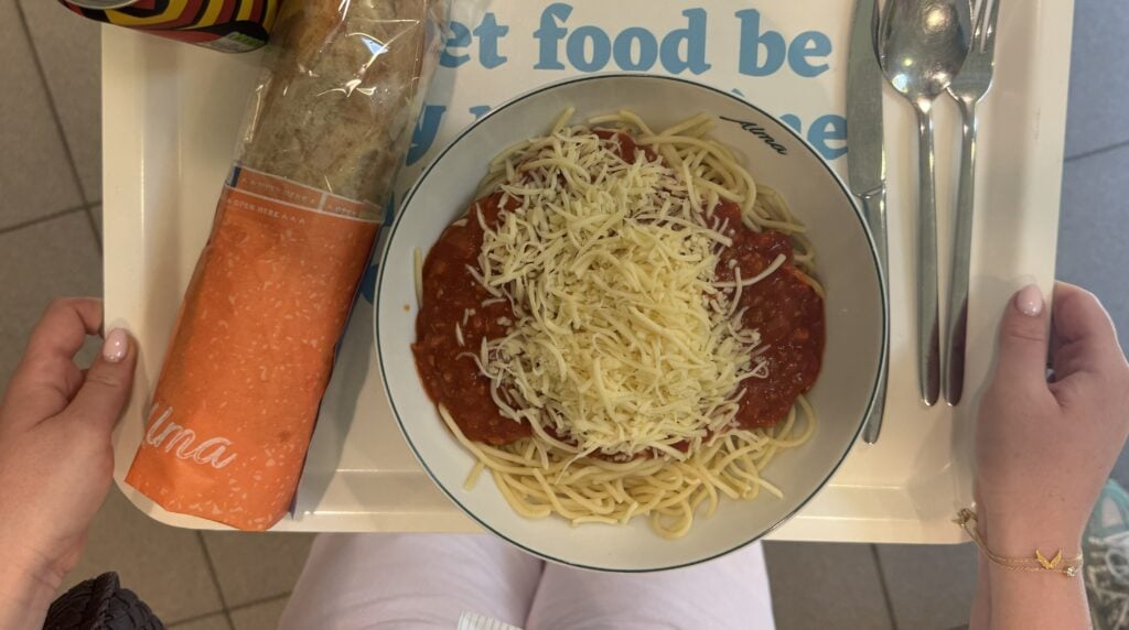 Iemand houdt een dienblad vast met daarop een kom spaghetti met kaas, een ingepakt stokbrood, een lepel en een vork – een klassiek campusgerecht, getest aan de KULeuven.