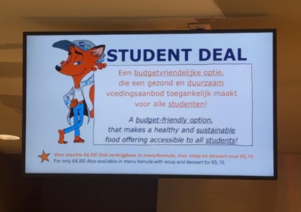 Op een digitaal scherm wordt een maaltijdaanbieding voor studenten getoond met een cartoonvos, waarmee zowel in het Nederlands als in het Engels betaalbare, gezonde en duurzame maaltijdopties worden gepromoot, met prijzen van € 4,50 en € 5,10. Dit campusvoedselconcept, dat is getest op de VUB in Brussel, zorgt ervoor dat studenten kunnen genieten van heerlijke maaltijden die zowel betaalbaar als milieuvriendelijk zijn.