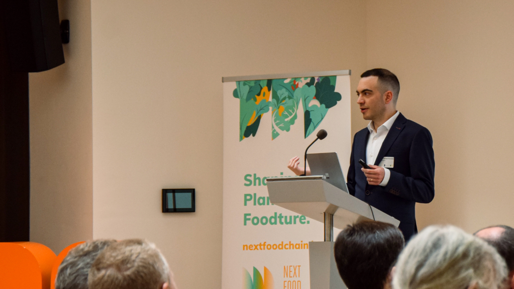 Een man in pak houdt een toespraak vanaf een podium voor een spandoek met de titel Shaping Plant-Based Foodtures.