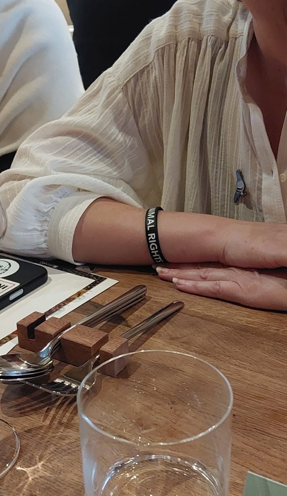 Iemand met een zwarte polsband waarop ANIMAL RIGHTS staat, zittend aan een houten tafel met bestek.
