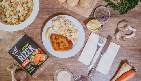 Na dřevěném stole leží talíř řízku s bramborovým salátem a zeleninou s citronem, obklopený brambory, mrkví, houbami, balenými veganskými řízky, nádobím, ubrousky a miskami na koření.