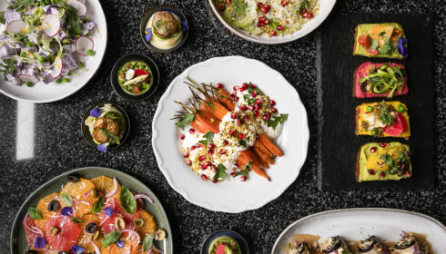 Pestrá nabídka barevných vegetariánských pokrmů, včetně salátů, pečených mrkví, dipů a toastů s zeleninovou oblohou, naservírovaná na černé pracovní desce – ideální volba pro ty, kdo se řídí proteinovým plánem EU a hledají pestré rostlinné pokrmy.