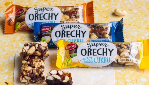 Na žlutém podkladu jsou rozmístěny tři tyčinky Emco Super Ořechy, z nichž dvě jsou bez obalu, s viditelnými ořechy a kousky čokolády. Tyto tyčinky, které zdůrazňují svou kvalitu, se pyšně chlubí veganským certifikátem od skupiny PEK. Jedna tyčinka bez obalu je umístěna v popředí.