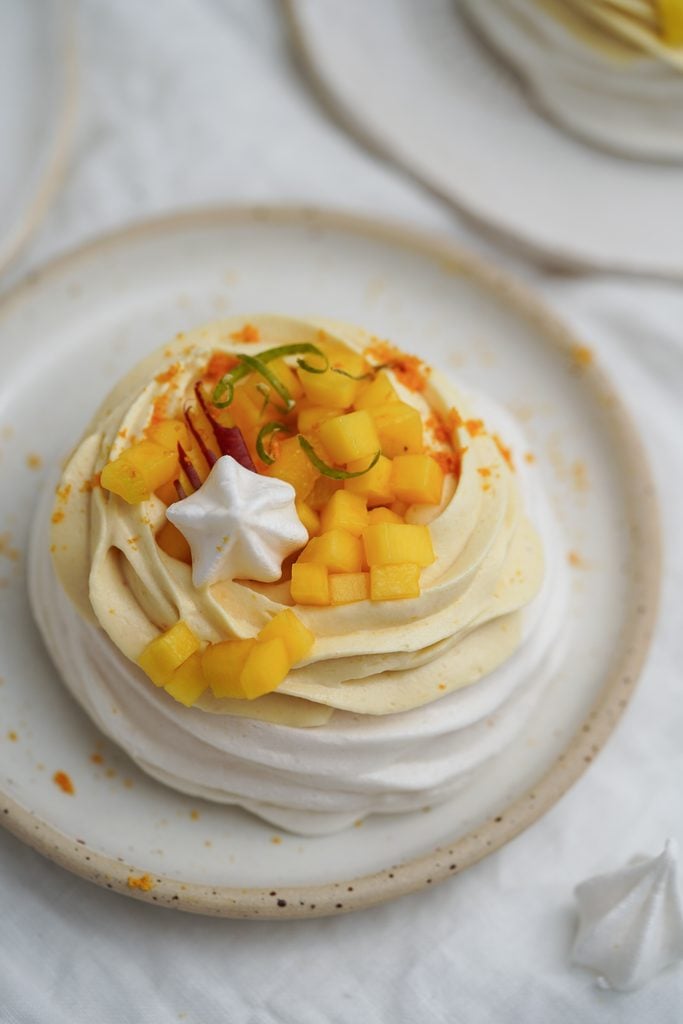 Na keramickém talíři leží dezert pavlova zdobený nakrájeným mangem, bílou meruňkovou hvězdou a citrusovou kůrou – elegantní příklad toho, jak může rostlinný catering na akci vnést do každé události rafinované chutě.