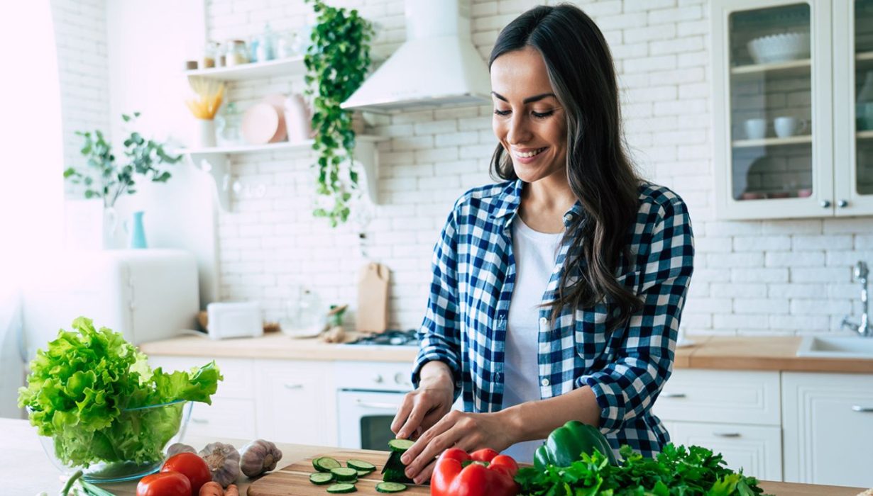 Eine Frau in einem karierten Hemd lächelt, während sie in einer hellen Küche, umgeben von frischem Gemüse wie Salat, Tomaten und Paprika auf einer Holztheke, Gurken schneidet.