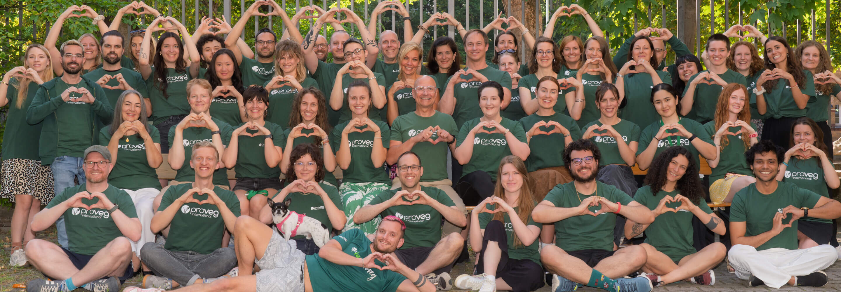 Eine große Gruppe von Menschen in grünen T-Shirts sitzt und steht gemeinsam im Freien, formt mit den Händen Herzchen und lächelt in die Kamera.