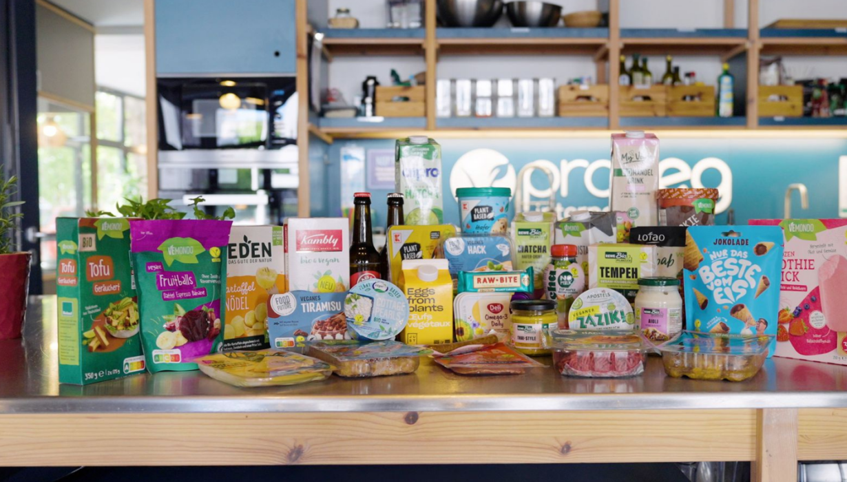 Eine Auswahl an verpackten veganen Lebensmitteln, ausgestellt auf einer Küchenarbeitsplatte, im Hintergrund Regale, Pflanzen und Küchenutensilien.