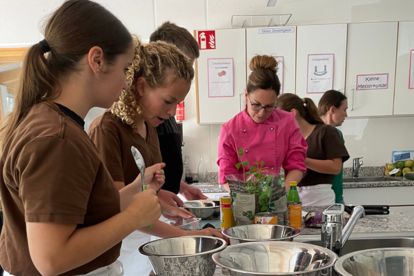 Vier Personen bereiten gemeinsam in einer Küche Essen zu, wobei sie Metallrührschüsseln und verschiedene Zutaten verwenden; eine Person in einer rosa Kochjacke beaufsichtigt sie dabei.