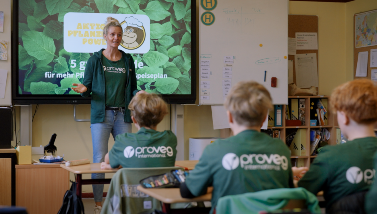 Eine Frau steht vor einer Klasse und hält den Schülern einen Vortrag. Auf der Leinwand hinter ihr ist ein Projekt zum Thema pflanzliche Ernährung zu sehen. Die Schüler tragen grüne T-Shirts mit dem ProVeg International-Logo.