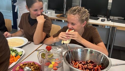 Zwei Mädchen in T-Shirts sitzen an einem Tisch mit Salat, Gemüse und Obstwasser; das eine Mädchen isst einen Wrap, während das andere sie anlächelt.