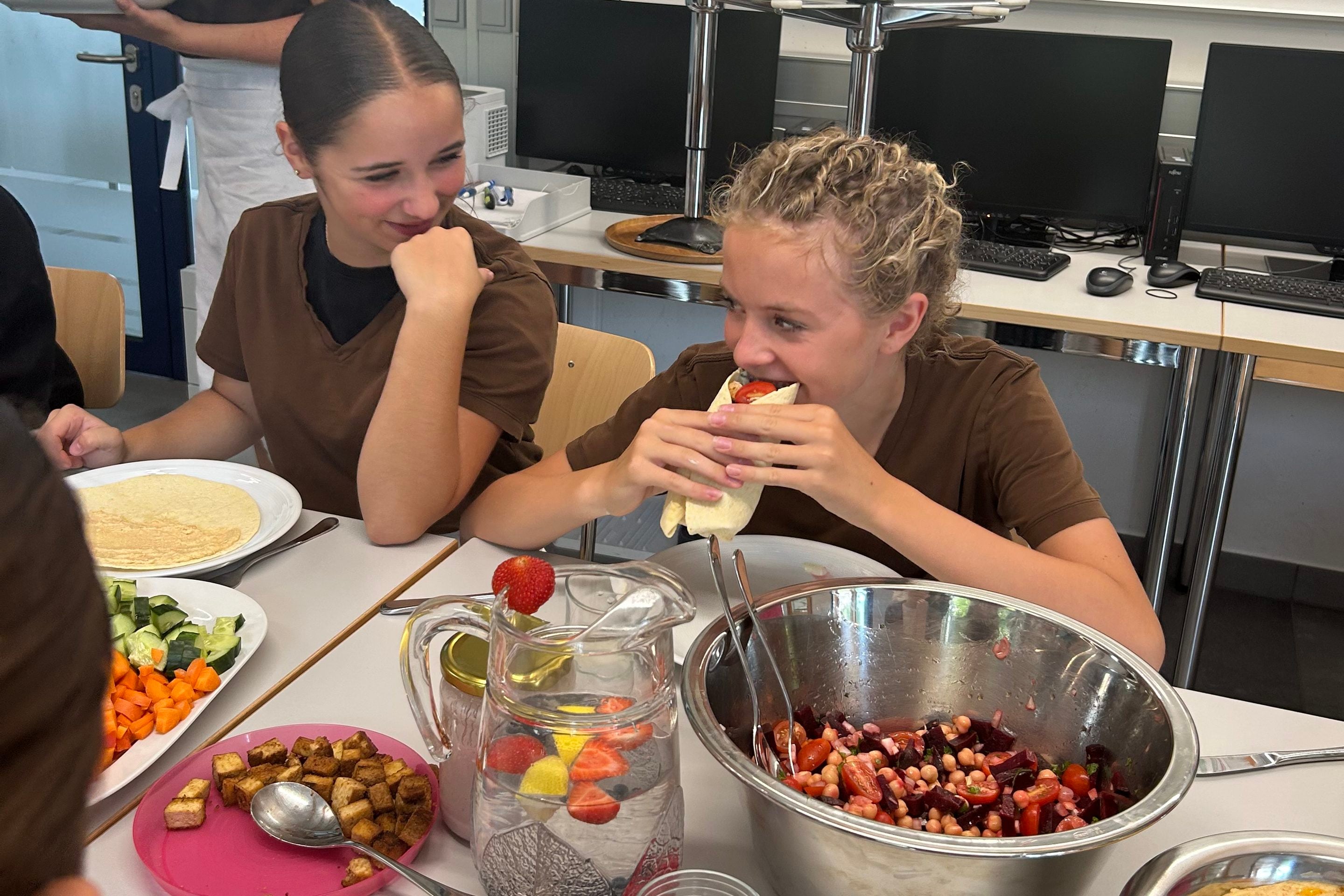 Zwei Mädchen in T-Shirts sitzen an einem Tisch mit Salat, Gemüse und Obstwasser; das eine Mädchen isst einen Wrap, während das andere sie anlächelt.
