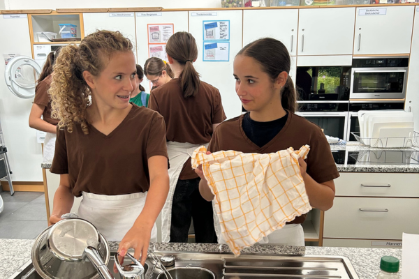 Zwei Mädchen in braunen Hemden und weißen Schürzen spülen und trocknen Geschirr an einem Spülbecken, während im Hintergrund drei weitere Personen arbeiten.