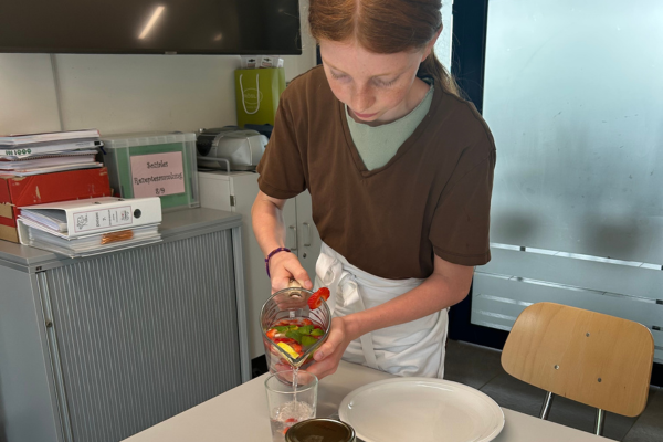 Eine Person gießt Wasser aus einem Glaskrug mit Obst in ein Glas in einer Küche oder einem Essbereich, neben einem Tisch und Büromaterial.