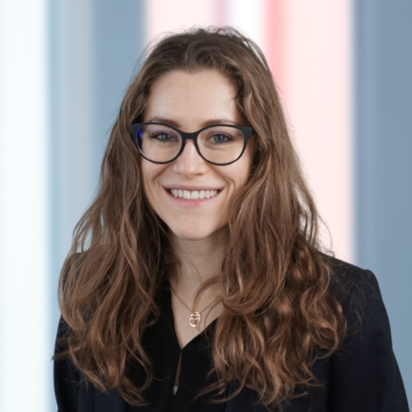 Eine Person mit langen braunen Haaren und Brille lächelt in die Kamera; sie trägt einen dunklen Blazer und eine Halskette, während im Hintergrund verschwommene blaue und rosa vertikale Streifen zu sehen sind – ein Bild, das die professionelle, einladende Atmosphäre einfängt, die oft mit BKK ProVita in Verbindung gebracht wird.