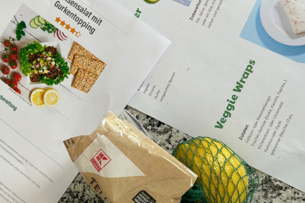 Auf einer grauen Arbeitsplatte liegen ausgedruckte Rezepte und Zutaten, darunter eine Packung Tee und eine in einem Netz verpackte Zitrone.