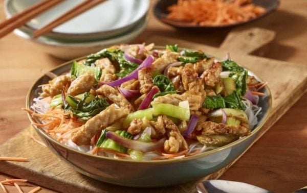 Un tazón de fideos de arroz cubiertos con tofu salteado, cebollas rojas en rodajas, pak choi, zanahorias y pepinos se encuentra sobre una tabla de madera, con zanahorias ralladas y platos en el fondo.