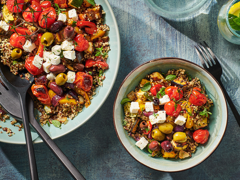 Dos tazones de ensalada mediterránea de quinoa con tomates cherry, verduras asadas, aceitunas y queso feta. Junto a los tazones hay un tenedor y un cuchillo, y al fondo, un vaso de agua con limón.