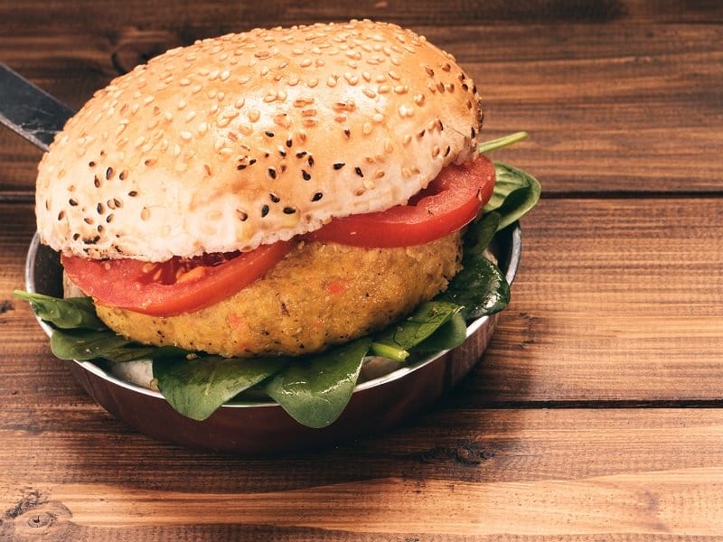 Un primer plano de una hamburguesa vegetariana con hojas de espinaca, rodajas de tomate y un pan con semillas de sésamo, servida en un plato redondo pequeño sobre una superficie de madera.