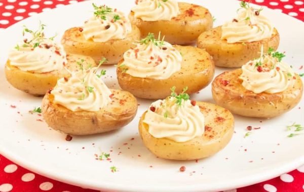 Un plato blanco contiene pequeñas mitades de papas horneadas cubiertas con crema en espiral y adornadas con hierbas y especias, sobre un mantel de lunares rojos y blancos.