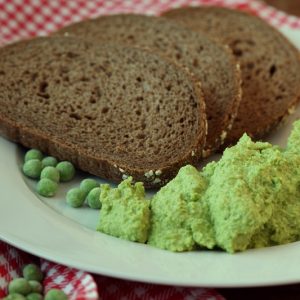 Se sirven tres rebanadas de pan de centeno oscuro en un plato blanco con una bola de pasta de guisantes y unos cuantos guisantes enteros. Debajo del plato se ve un mantel rojo a cuadros.