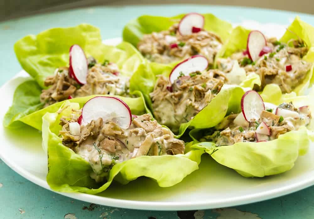 Un plato de lechuga rellena de ensalada cremosa de pollo, adornado con rábanos en rodajas finas y hierbas frescas, dispuestos cuidadosamente en una bandeja blanca.