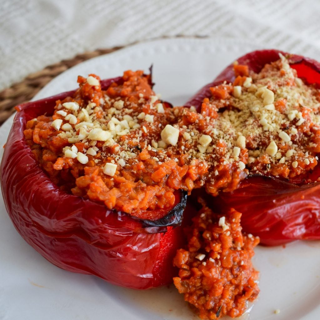 Un pimiento rojo asado cortado por la mitad y relleno con una mezcla de arroz a base de tomate, cubierto con nueces trituradas, servido en un plato blanco.