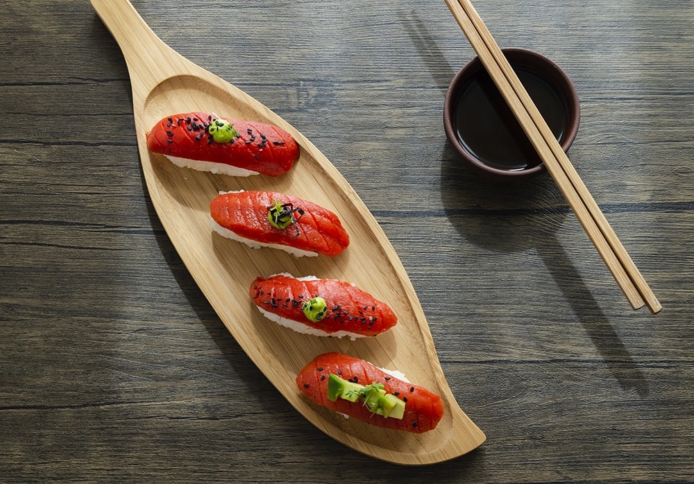 Cuatro piezas de sushi nigiri de atún están dispuestas en un plato de madera con forma de hoja, adornadas con cebollas verdes y sésamo negro, con un recipiente con salsa de soja y palillos al lado, en una mesa de madera.