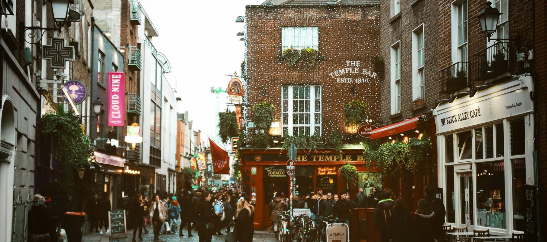 Una bulliciosa calle adoquinada de Dublín, Irlanda, con multitudes caminando junto al pub The Temple Bar, rodeado de edificios históricos de ladrillo, vegetación y carteles de cafeterías.