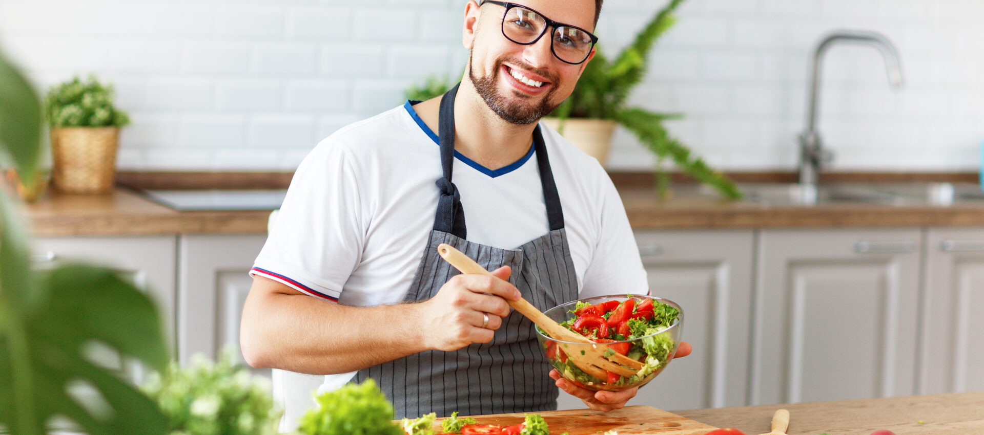 Un hombre sonriente con gafas y delantal mezcla una ensalada fresca en un bol de cristal en una cocina luminosa, rodeado de verduras y plantas.