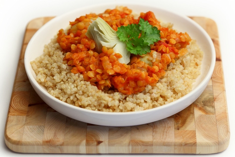 Un bol de quinoa cubierto con un guiso de lentejas a base de tomate, adornado con un corazón de alcachofa y cilantro fresco, colocado sobre una tabla de cortar de madera.