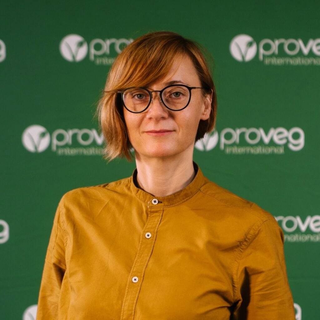 Osoba o krótkich, jasnobrązowych włosach i w okularach, ubrana w koszulę w kolorze musztardowej żółci, stoi przed zielonym tłem z logo firmy Proveg International.