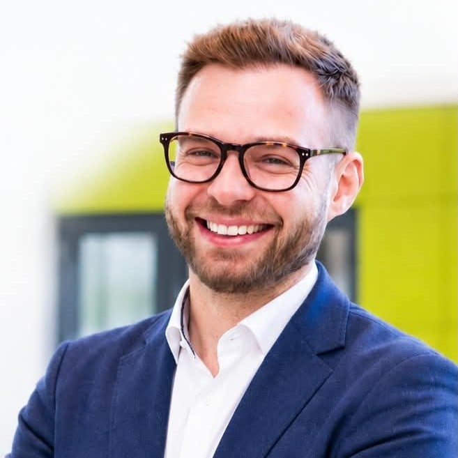 Mężczyzna w okularach i z brodą, ubrany w niebieską marynarkę i białą koszulę, uśmiecha się w jasno oświetlonym wnętrzu, uosabiając innowacyjnego ducha Szkoły Przyszłości.