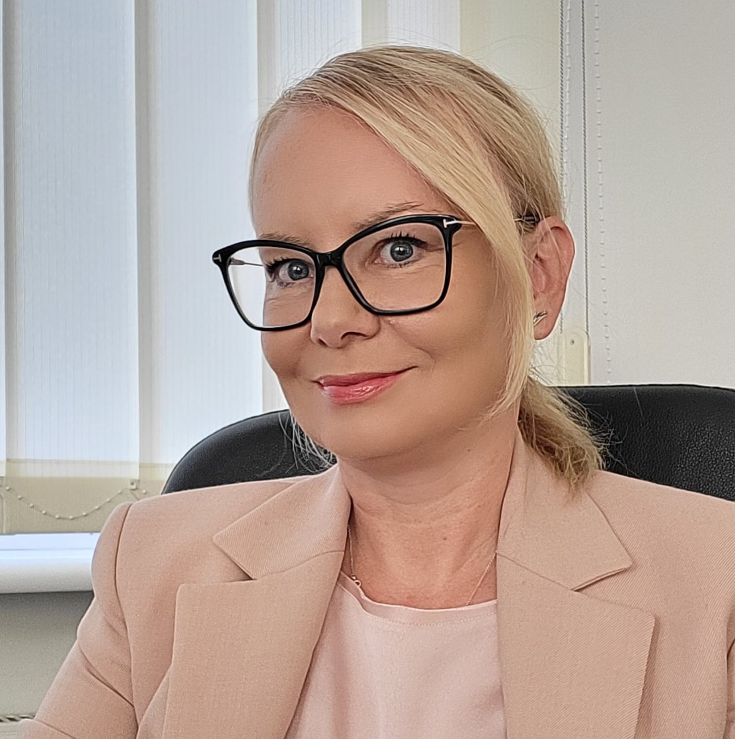 Kobieta o blond włosach i w okularach siedzi na krześle biurowym, ubrana w jasnoróżową marynarkę i lekko się uśmiecha. Okno za nią zasłaniają żaluzje pionowe, tworząc profesjonalną atmosferę, która odzwierciedla innowacyjnego ducha Szkoły Przyszłości.
