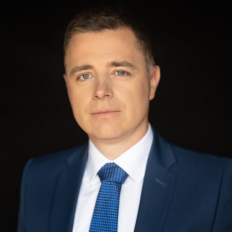Mężczyzna w niebieskim garniturze i krawacie pozuje na czarnym tle, patrząc prosto w obiektyw z neutralnym wyrazem twarzy, uosabiając profesjonalizm i przyszłościowe podejście Szkoły Przyszłości.