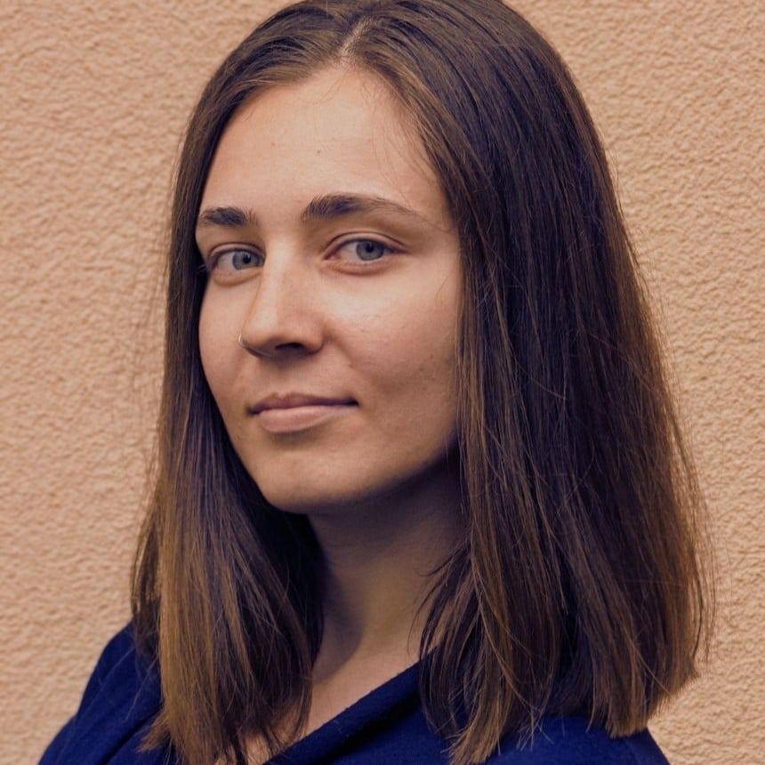 Kobieta o prostych brązowych włosach i niebieskich oczach patrzy w obiektyw, stojąc przed fakturowaną beżową ścianą. Ma na sobie ciemną bluzkę i zachowuje neutralny wyraz twarzy, uosabiając spokojną pewność siebie kojarzoną ze Szkołą Przyszłości.