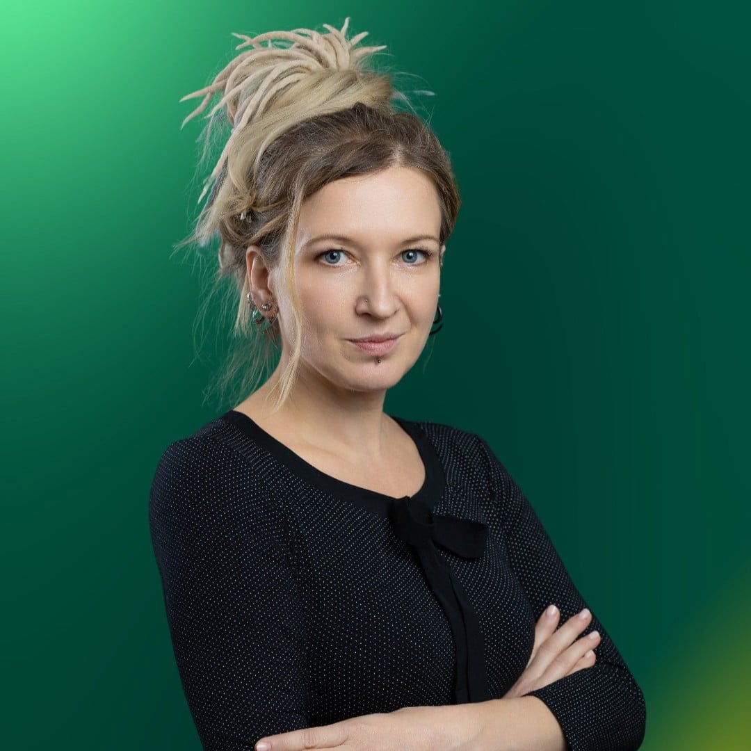 Kobieta o blond włosach upiętych w kok, ubrana w czarną bluzkę o fakturze, stoi ze skrzyżowanymi rękami na tle zielonego gradientu, uosabiając pewność siebie i wizję Szkoły Przyszłości.